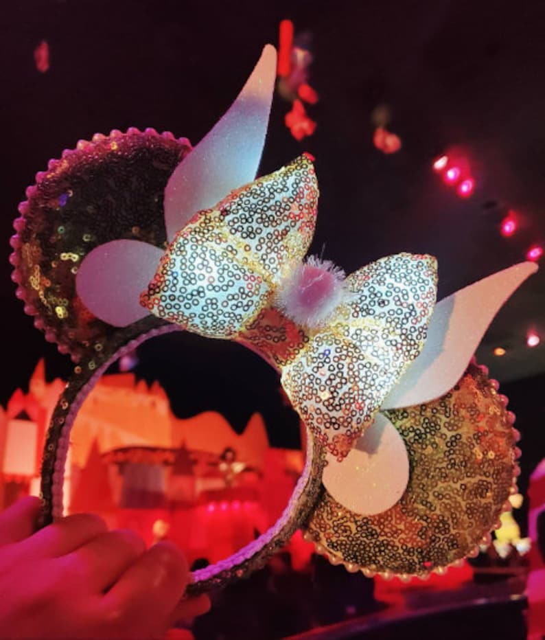 K&ouml;nnte beinhalten: Goldene und wei&szlig;e Pailletten-Minnie-Maus-Ohren mit einer gro&szlig;en wei&szlig;en Schleife und wei&szlig;en Hasenohren. Die Ohren sind mit kleinen, glitzernden Pailletten verziert.