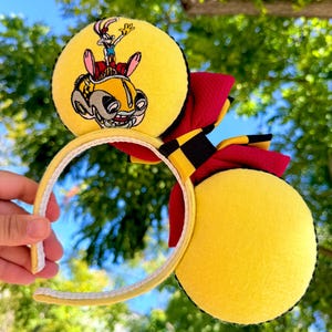 May include: Yellow mouse ears with a cartoon car and character design. The ears have a red and black bow and a yellow headband. The design features a cartoon character in a yellow car.