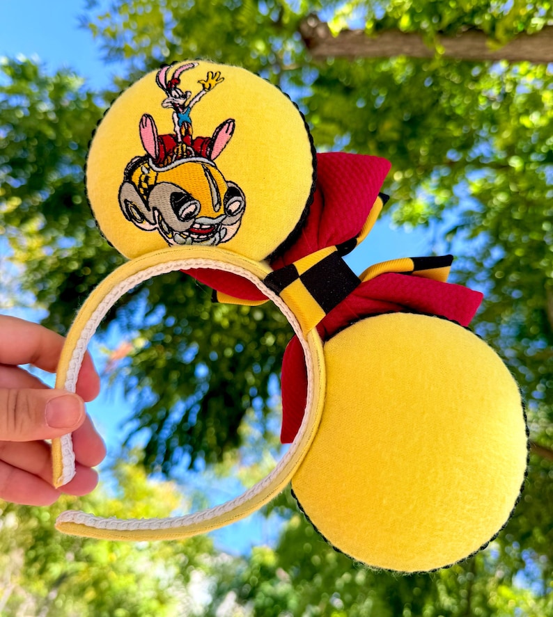 May include: Yellow mouse ears with a cartoon car and character design. The ears have a red and black bow and a yellow headband. The design features a cartoon character in a yellow car.