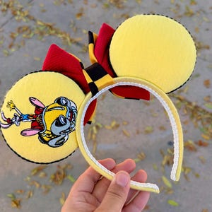 May include: Yellow mouse ear headband with a red and black bow. One ear features a cartoon character illustration. The headband is yellow with a white trim. The ears are yellow with black stitching.