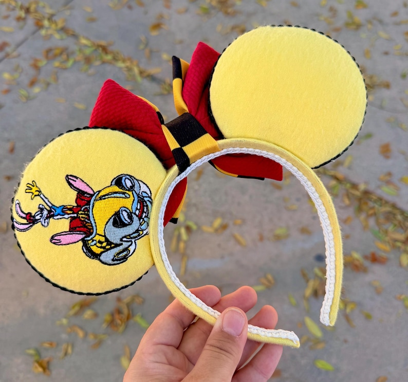 May include: Yellow mouse ear headband with a red and black bow. One ear features a cartoon character illustration. The headband is yellow with a white trim. The ears are yellow with black stitching.