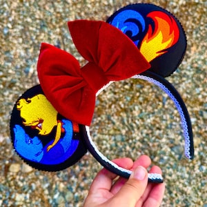 May include: A pair of black mouse ears with a red bow. One ear features an embroidered image of a blue and yellow flame, while the other ear features an embroidered image of a blue and yellow character.