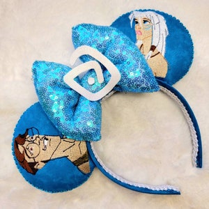 May include: Blue and white Disney-themed mouse ears with a sequined bow and embroidered images of characters from the movie 'The Princess and the Frog'.