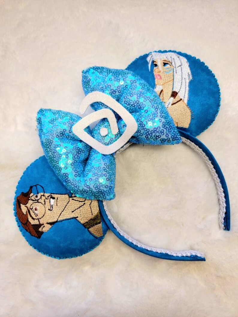 May include: Blue and white Disney-themed mouse ears with a sequined bow and embroidered images of characters from the movie 'The Princess and the Frog'.