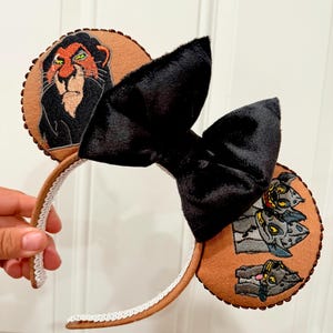 May include: Brown mouse ear headband with embroidered images of Scar and hyenas. The headband features a large black bow and a white lace trim along the band. The ears are a light brown color.