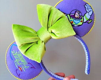 Könnte beinhalten: Lila und grüne Minnie Mouse Ohren mit einer grünen Samtschleife und einem gestickten Bild von Buzz Lightyear aus Toy Story. Die Ohren haben einen gelben Rand und das Stirnband ist weiß mit einem lila Rand.
