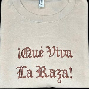 May include: A folded, beige t-shirt with the text "¡Qué Viva La Raza!" embroidered in brown. The shirt is made of a soft material and has a crew neck. The tag "American Apparel" is visible at the top.