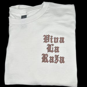 May include: White t-shirt with the phrase "Viva La Raza" printed in brown script.