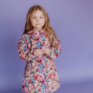 May include: A young girl wearing a floral print jacket with long sleeves and a white background. She is wearing blue jeans and white sneakers. The girl is standing on a purple background.