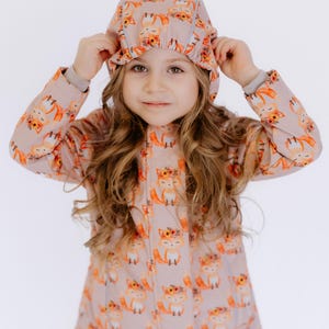 May include: A young girl wearing a pink and orange jacket with a fox print. The jacket has a hood and a zipper closure. The girl is also wearing a matching hat with a fox print.