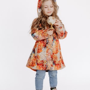 Puede incluir: Una niña pequeña con un impermeable con capucha de colores con un patrón de burbujas, jeans azules y botas negras. Está sosteniendo un pequeño globo terráqueo en sus manos.