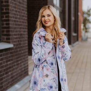Può includere: Una donna indossa un impermeabile bianco con stampa floreale. L'impermeabile ha un cappuccio e una chiusura a zip. La donna è in piedi davanti a un muro di mattoni.