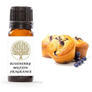 Op de afbeelding: Een klein amberkleurig glazen flesje met een zwarte dop en een wit etiket. Het etiket heeft een boomontwerp en de tekst "Blueberry Muffin Fragrance". Het flesje staat naast een foto van bosbessenmuffins.