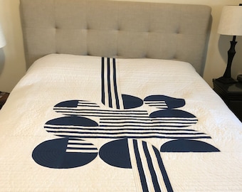 Puede incluir: Una colcha a rayas blancas y azules con un diseño geométrico de círculos azul marino. La colcha está sobre una cama con una cabecera gris claro.