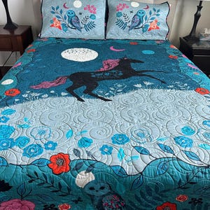 May include: A quilt and two matching pillow shams featuring a dark teal background with a black unicorn, pink mane and tail, white moon, and owls. The design includes floral patterns in red, pink, and blue, with swirling quilted stitching throughout.