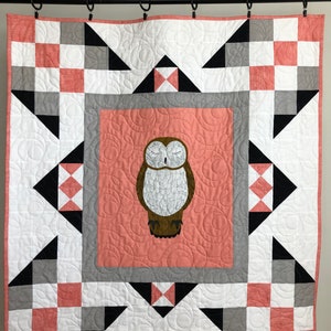 May include: A white, black, and pink quilt with a brown owl design in the center. The quilt is hanging on a black curtain rod.