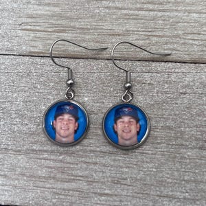 Könnte beinhalten: Ein Paar runde, silberfarbene Ohrringe. Jeder Ohrring zeigt ein kreisförmiges Bild einer Person, die eine Baseballkappe mit einem blau-weißen Logo trägt. Der Hintergrund des Bildes ist blau. Die Ohrringe hängen an einem silbernen Haken.