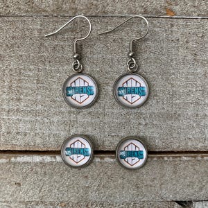 May include: A pair of silver-toned drop earrings and a pair of matching stud earrings. Each piece features a circular glass cabochon with a white background, a geometric orange outline, and the teal text SIRENS printed across the centre.