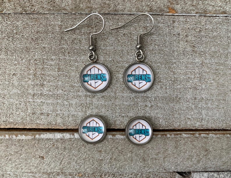 May include: A pair of silver-toned drop earrings and a pair of matching stud earrings. Each piece features a circular glass cabochon with a white background, a geometric orange outline, and the teal text SIRENS printed across the centre.