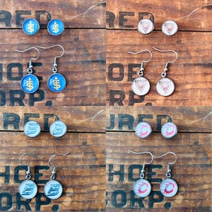 May include: A collection of four pairs of circular earrings featuring various designs including blue and gold symbols, white and red geometric logos, and silver or red circular patterns, all suspended from silver-toned metal earring hooks.