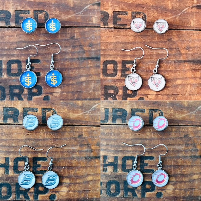 May include: A collection of four pairs of circular earrings featuring various designs including blue and gold symbols, white and red geometric logos, and silver or red circular patterns, all suspended from silver-toned metal earring hooks.