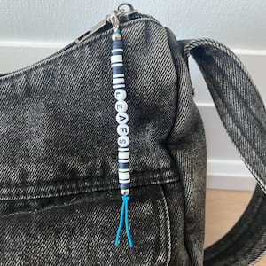 Op de afbeelding: Een zwarte acid-wash denim schoudertas met een rits-bedel. De bedel bestaat uit afwisselend donkerblauwe en witte cilindrische kralen met witte ronde letterkralen die LEAFS spellen, afgewerkt met een felblauw koordkwastje aan het uiteinde.