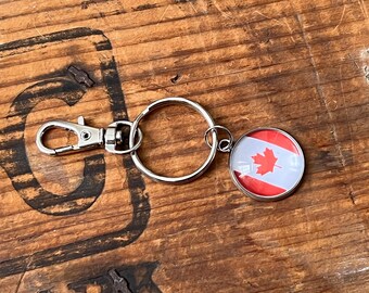 May include: A silver-colored keychain with a round charm featuring the Canadian flag. The charm has a red maple leaf on a white background, with red stripes on the top and bottom. The keychain includes a clasp and a ring.