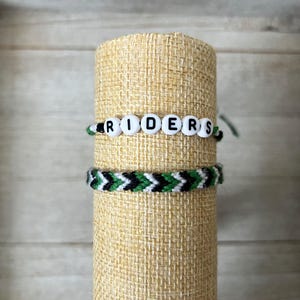 May include: Two handmade bracelets. The top bracelet has white letter beads spelling "RIDERS" on a green and white string. The bottom bracelet is a chevron pattern in green, black, and white.