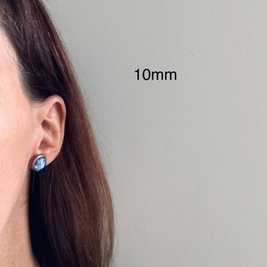 May include: A round stud earring with a blue and white marbled pattern set in a silver-toned metal frame. The earring measures 10mm in diameter and is positioned on an earlobe, showcasing the circular design and the specific size measurement.