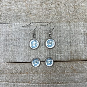 May include: A pair of silver-toned drop earrings and a pair of matching stud earrings, each featuring a circular glass cabochon with a light blue letter S and the word TORRENT printed in white text against a white background.