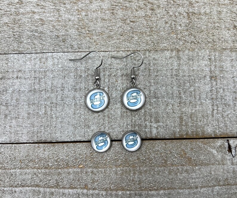 May include: A pair of silver-toned drop earrings and a pair of matching stud earrings, each featuring a circular glass cabochon with a light blue letter S and the word TORRENT printed in white text against a white background.