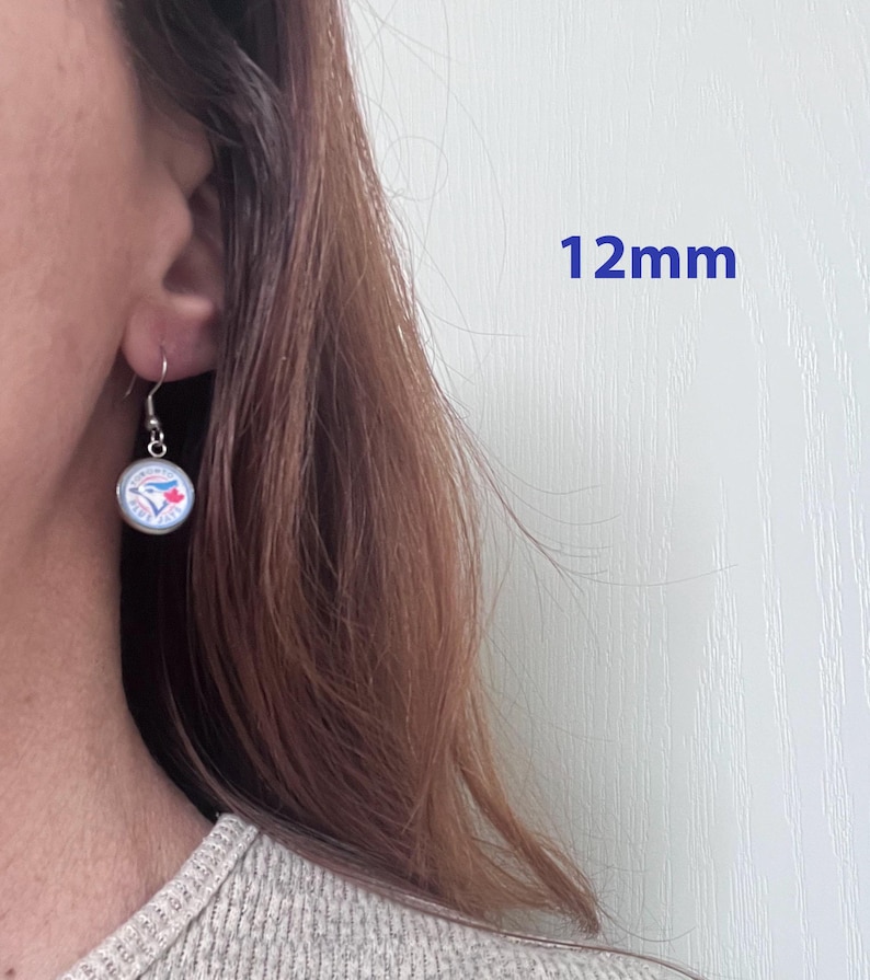 May include: A silver-toned drop earring featuring a circular charm with the Toronto Blue Jays logo, which includes a blue bird and a red maple leaf. The charm has a diameter of 12mm and hangs from a simple silver-toned hook wire.