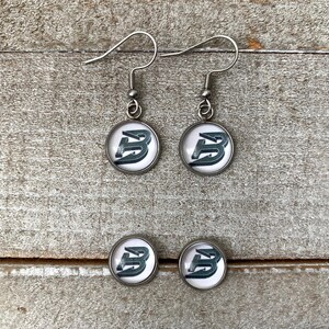 May include: A pair of silver-toned drop earrings and a pair of matching stud earrings, each featuring a circular setting with a white background and a dark, stylised letter B graphic printed in the centre, complete with silver-toned metal hooks.
