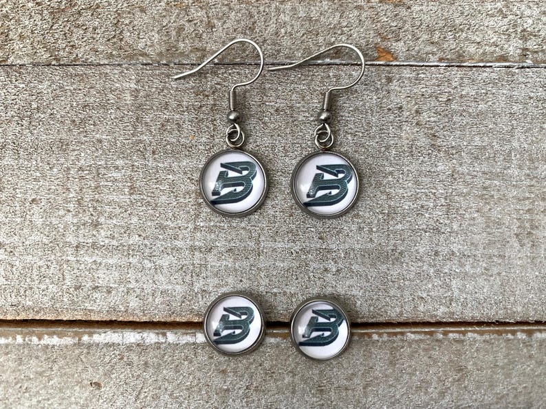 May include: A pair of silver-toned drop earrings and a pair of matching stud earrings, each featuring a circular setting with a white background and a dark, stylised letter B graphic printed in the centre, complete with silver-toned metal hooks.