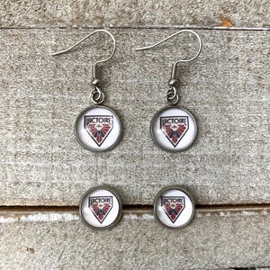 May include: A pair of silver-toned drop earrings and a pair of matching stud earrings. Each piece features a circular glass cabochon with a white background, a red and blue shield-shaped emblem, and the word VICTOIRE printed in black text.