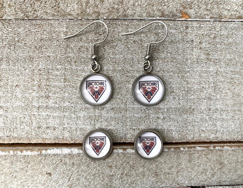 May include: A pair of silver-toned drop earrings and a pair of matching stud earrings. Each piece features a circular glass cabochon with a white background, a red and blue shield-shaped emblem, and the word VICTOIRE printed in black text.