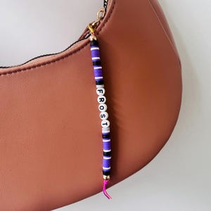 May include: A brown handbag features a dangling charm made of purple, black, and white disc beads. The charm includes white letter beads spelling out the word FROST, with a small gold-tone bead at the top and a bright pink string at the bottom.