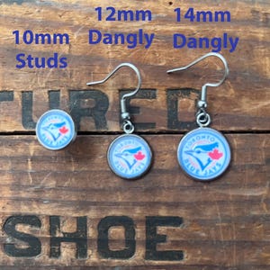 May include: A set of Toronto Blue Jays earrings featuring one pair of 10mm studs, one pair of 12mm dangly earrings, and one pair of 14mm dangly earrings. Each earring displays the blue jay logo with a red maple leaf on a white background.