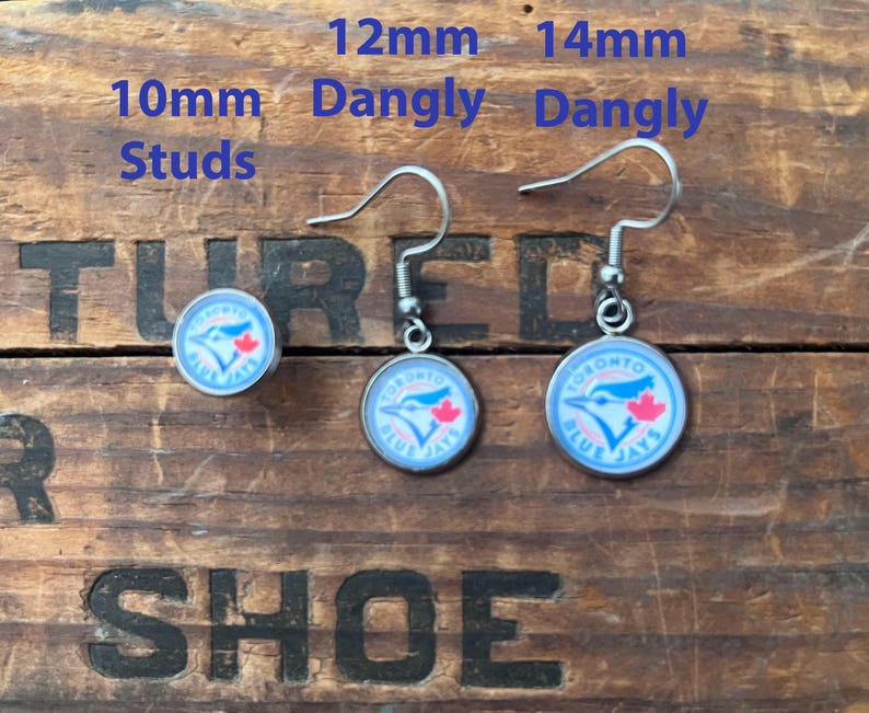 May include: A set of Toronto Blue Jays earrings featuring one pair of 10mm studs, one pair of 12mm dangly earrings, and one pair of 14mm dangly earrings. Each earring displays the blue jay logo with a red maple leaf on a white background.