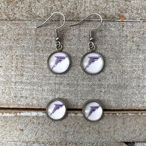 May include: A pair of silver-toned drop earrings and a pair of matching stud earrings. Each piece features a circular setting with a white background and a purple letter F graphic with a lightning bolt design integrated into the letter's shape.