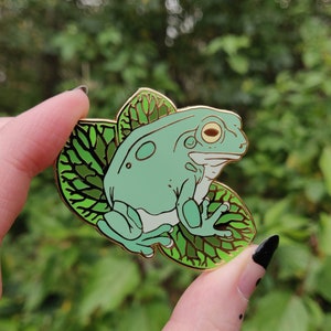 May include: A light green frog enamel pin with gold-colored accents, perched on a green leaf. The frog has large, round eyes and is set against a blurred green background. The pin is held in a hand.