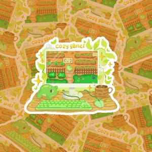 May include: A sticker featuring a pixelated illustration of a computer screen displaying a cosy garden scene. The text "Cozy Gamer" is displayed on the sticker. The computer has a green keyboard and a mouse. The sticker is surrounded by other stickers with the same design.