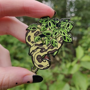 Peut inclure: Une épingle en émail dorée représentant un serpent vert enroulé autour d'une plante feuillue. Le serpent est jaune clair avec des marques brunes. La plante a des feuilles vertes avec des taches blanches.