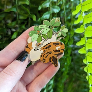 Op de afbeelding: Een gouden emaille pin met een bruine en witte slang die om een ​​groene plant gekruld is. De slang heeft een witte buik en bruine markeringen. De plant heeft groene bladeren en een bruine stengel.