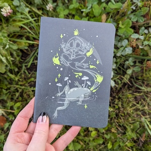 Puede incluir: Una libreta negra con una ilustración blanca y amarilla de un esqueleto de rana con ojos brillantes y una rana sentada en un hongo. El esqueleto de rana está rodeado de estrellas y humo amarillo brillante.