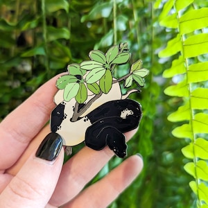 May include: Black and white enamel pin of a snake with a green plant growing out of its head.