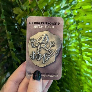 May include: A brown card with the text "FROG&TREEHOUSE MAY24 FOSSIL" and a silver frog skeleton fossil pin.