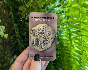 Op de afbeelding: Een bruine kaart met de tekst "FROG&TREEHOUSE MAY24 FOSSIL" en een zilveren speld met een fossiel skelet van een kikker.