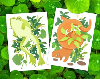 May include: Two illustrations on white cards, featuring a green frog and an orange salamander, both surrounded by green leaves. The frog is perched on a branch, while the salamander is curled around a plant.