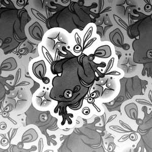 May include: A collection of grayscale stickers featuring a stylized frog design. The frog is depicted with large eyes, and is surrounded by leaves and stars. The stickers have a white border and are scattered across the image.